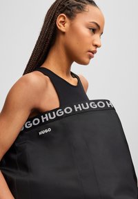 Borsa tote nera con finitura lucida, dotata di una spallina larga con "HUGO" in lettere grigie. Design semplice, forma rettangolare, interno spazioso.