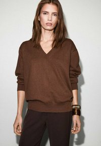 Suéter marrón de cuello en V con un ajuste holgado y puños acanalados, combinado con pantalones oscuros. Los accesorios incluyen una pulsera grande y texturizada.