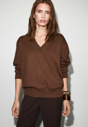 Pull marron à col en V avec une coupe oversize et des poignets côtelés, associé à un pantalon sombre. Les accessoires comprennent un grand bracelet texturé.