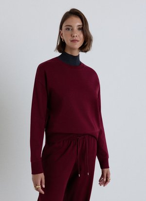 Donna con capelli castani corti che indossa una felpa rosso scuro e pantaloni abbinati, in piedi davanti a uno sfondo grigio chiaro uniforme.
