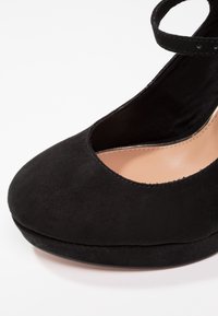 Chaussure pour femme en daim noir à bout fermé avec bride à la cheville et semelle intérieure beige, présentée en gros plan sur un fond blanc.