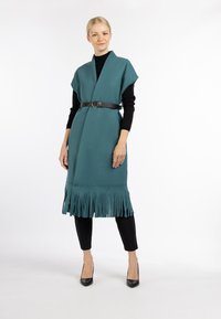 Teal fringed wikkeljurk met een diepe V-hals, ceintuur in de taille met een zwarte leren riem, gedragen over zwarte aansluitende broeken en hakken.
