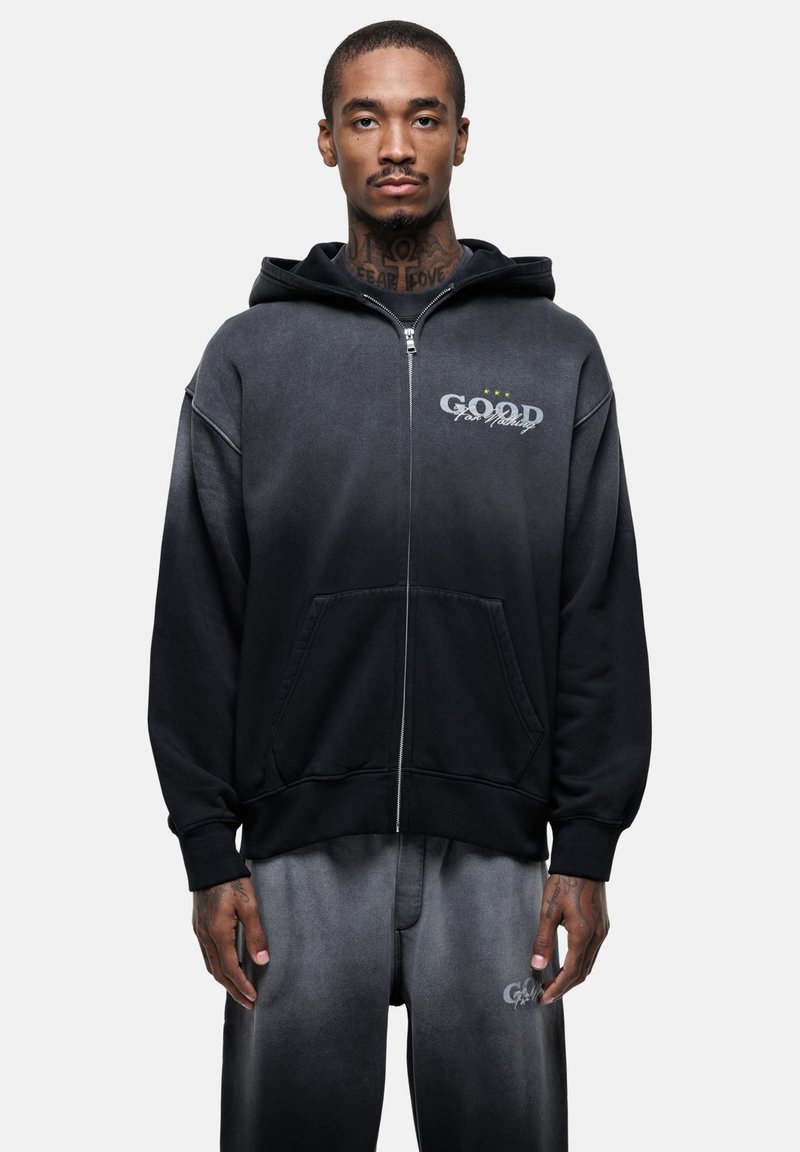 Hoodie noir dégradé à zip avec une poche avant et le mot "GOOD" imprimé en gris. Tissu doux avec une coupe décontractée, équipé d'une capuche à cordon.