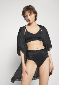 Frau trägt schwarzen Bralette, High-Waist-Slips mit transparenten Einsätzen und einen schwarzen Kimono-Morgenmantel mit Spitzenbesatz, steht vor einem einfarbigen Hintergrund.