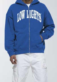 Felpa blu con zip e grafica bianca "LOW LIGHTS" sul petto. Presenta una tasca frontale e un cappuccio foderato. Pantaloni cargo bianchi visibili sotto.