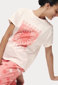 Kurzarmpullover aus cremefarbenem Baumwollstoff mit einem pinken Sonnenmotiv und dem Text "BONJOUR SOLEIL", kombiniert mit pinken Shorts, die mit Sonnenmustern verziert sind.