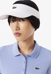 Visera blanca con una banda texturizada, logo de cocodrilo verde en la parte frontal, llevada con una camisa polo de color azul claro con cuello clásico y botones.