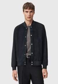 Giacca nera in suede con collo rotondo, chiusure a scatto e polsini a coste. Indossata sopra una maglietta grigia. Taglio dritto e design minimalista.