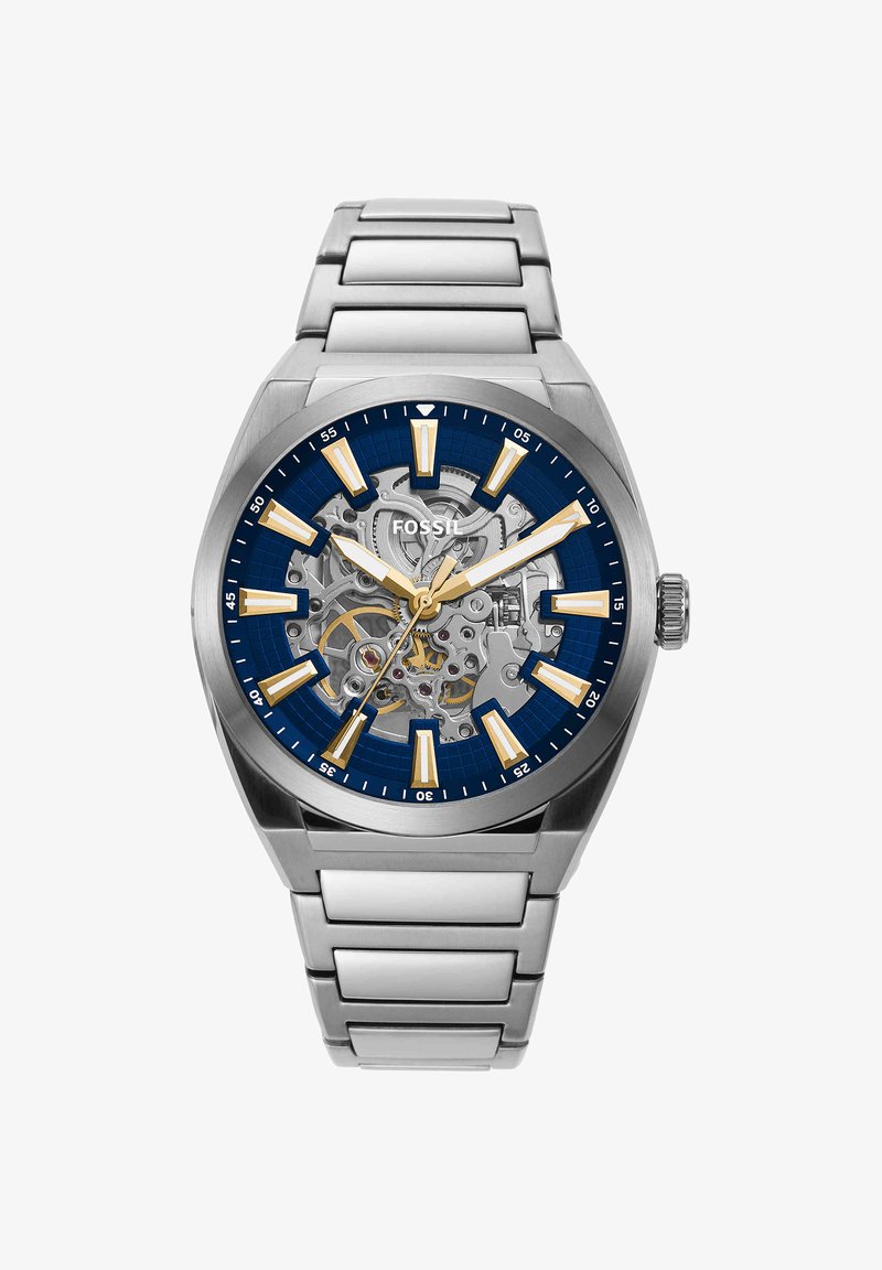Montre-bracelet Fossil argentée avec cadran squelette bleu, index dorés et bracelet à maillons en acier inoxydable, affichant les engrenages internes et l'heure.