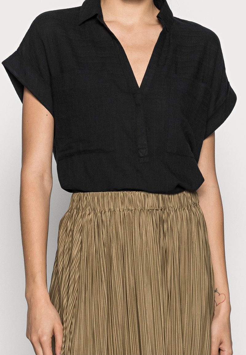 Svart kortärmad blus med V-ringning och framknäppning, i kombination med en veckad, beige kjol som har en texturerad tyg.