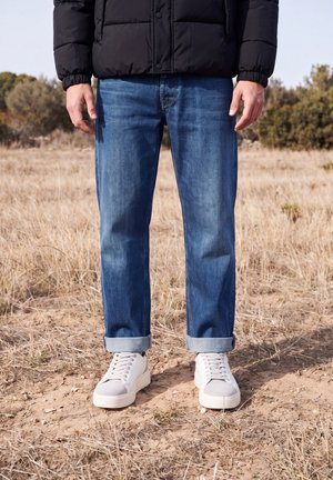 Une personne portant une doudoune noire, un jean bleu retroussé et des baskets blanches se tient dans un champ d’herbe sèche.