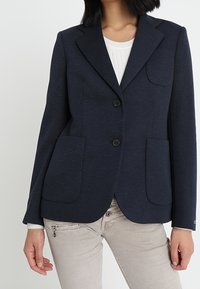 Blazer navy con un corte ajustado, solapas de muesca, dos bolsillos frontales y cierre de botón. Hecho de una textura de tela suave.