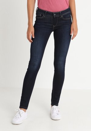 Vrouw die donkerblauwe skinny jeans, een roze top en witte sneakers draagt, staand tegen een effen lichte achtergrond.