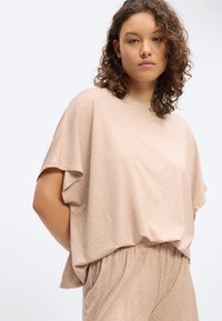 Γυναίκα με κατσαρά μαλλιά που φορά χαλαρό, μπεζ κοντομάνικο τοπ και ταιριαστό παντελόνι ψηλής μέσης, μπροστά σε απλό ανοιχτόχρωμο φόντο.