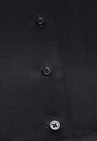 Chemise noire avec une texture lisse, dotée de quatre boutons noirs et d'un motif à côtes subtil. Boutons situés à mi-hauteur du tissu.