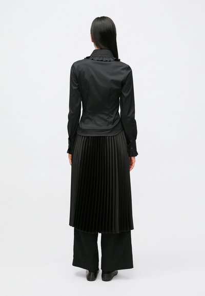 Melnā krekla ar garām piedurknēm un volānu apkakli, kopā ar melnu plisētu svārku. Apģērbam ir gludas līnijas un gluda faktūra.