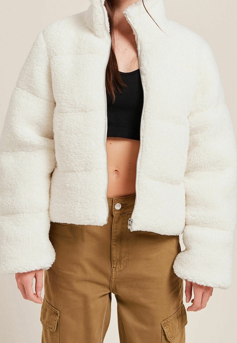 Chaqueta corta blanca y esponjosa con cremallera delantera, cuello alto y mangas anchas. La tela tiene una apariencia texturizada y aterciopelada.