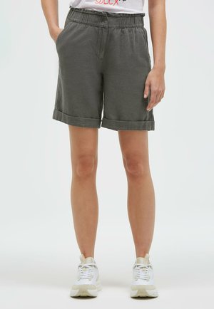 Person trägt olivgrüne Shorts mit umgeschlagenem Saum und elastischem Bund sowie weiße Sneaker, steht mit einer Hand in der Tasche.