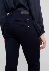 Person som bär mörkblå byxor och matchande skjorta, hand avslappnat vid sidan, synliga bakfickor med knapp och liten lädermärkeslapp.