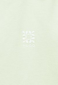 Λευκό ύφασμα με ένα διακριτικό ανάγλυφο λογότυπο που παρουσιάζει μια αφηρημένη συμμετρική μορφή λουλουδιού και τη λέξη «HUGO» από κάτω.