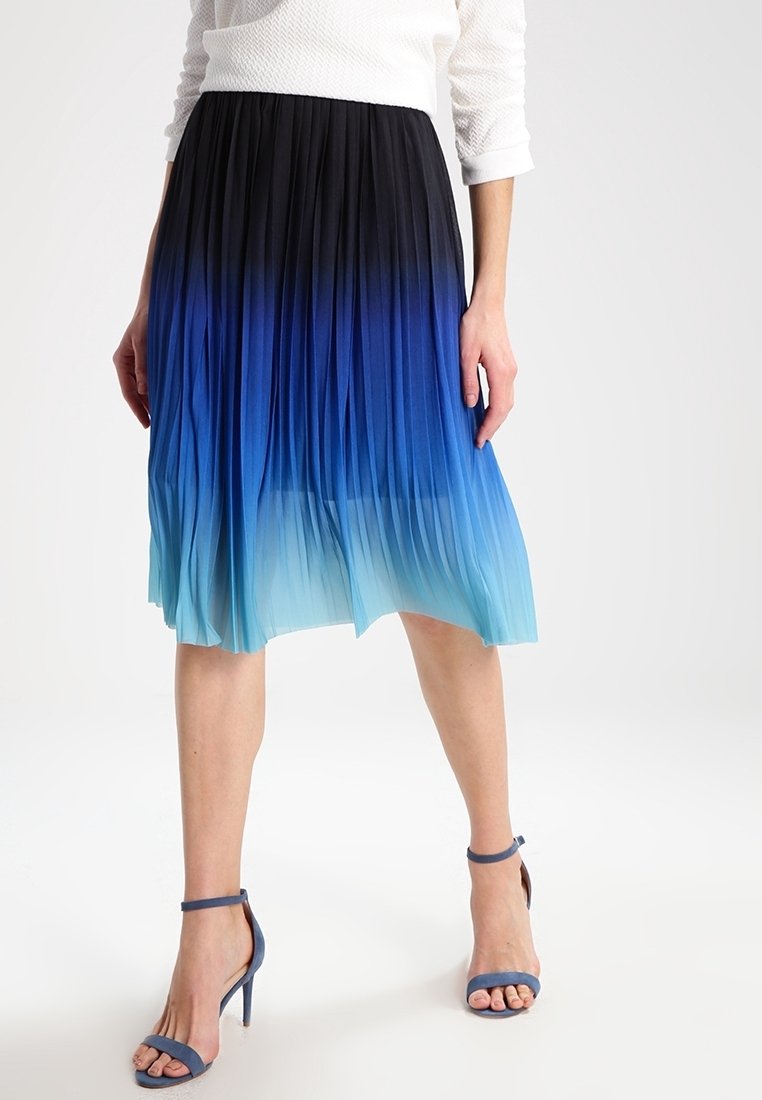 Femme portant une jupe midi plissée avec un dégradé du bleu au noir et des sandales à talons hauts bleues, debout devant un fond blanc.