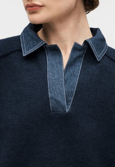 Gros plan d'une personne portant un haut bleu foncé avec un col ouvert à texture denim et des détails de couture clairs.