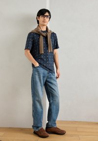 Ναυτική κοντομάνικη μπλούζα με πολύχρωμο λουλουδάτο σχέδιο, καφέ πουλόβερ ριγμένο γύρω από τον λαιμό, μπλε τζιν και καφέ παπούτσια.