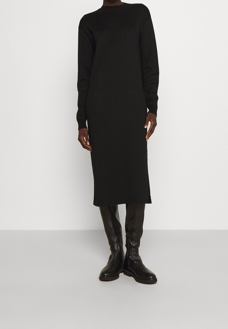 Personne portant une longue robe-pull noire et des bottes en cuir noires montant aux genoux, debout devant un fond blanc uni.