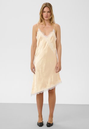 Femme blonde debout portant une robe nuisette en satin pêche pâle avec bordure en dentelle et des ballerines pointues noires sur un fond clair uni.