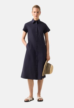 Kvinde iført en marineblå midi-kjole med knapper og korte ærmer, der holder en stribet halmhut og bærer beige sandaler.