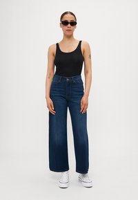 Mørkeblå vid jeans med høj talje og forlommer, parret med en sort tanktop og hvide sneakers, accentueret af mørke solbriller.
