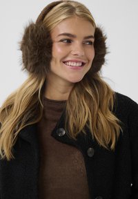 Rudos spalvos dirbtinės kailio ausinės su šokoladinės rudos spalvos audinio juosta, dėvimos ant ilgų blondinių plaukų, derinamos su tamsia striuke ir rudu megztiniu.