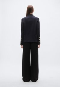 Zwart colbert met getailleerde pasvorm, revers met inkeping en gouden knopen op de mouwen, gecombineerd met zwarte broek met wijde pijpen. Soepele stoftextuur.