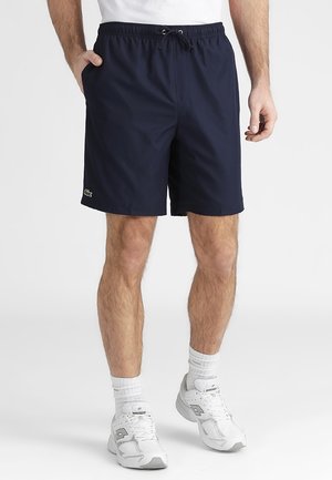 Hombre con pantalones cortos azul marino con cordón, calcetines blancos y zapatillas deportivas blancas de pie con una mano en el bolsillo.