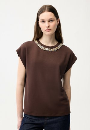 Jeune femme aux longs cheveux bruns portant un haut marron sans manches à décolleté orné et un jean bleu foncé, debout devant un fond uni.