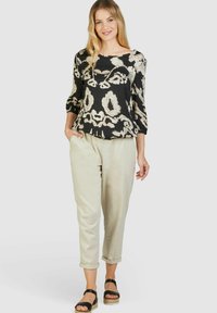 Schwarz-beige gemusterte Bluse mit Dreiviertelärmeln, kombiniert mit hellen, hochgekrempelten Hosen und schwarzen Sandalen.