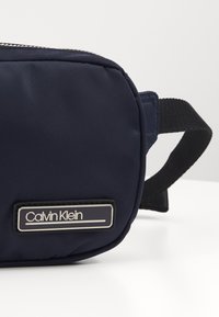 Granatowa torebka Calvin Klein z czarnym regulowanym paskiem i srebrnym zamkiem błyskawicznym, umieszczona na białej powierzchni na tle jasnoszarym.