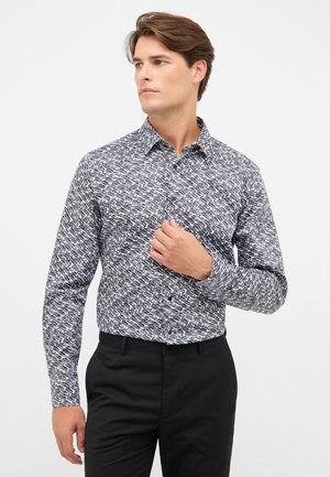 Man in zwarte broek en een overhemd met lange mouwen, voorzien van een zwart-wit geometrisch patroon, die het overhemd bij de borst aanpast en naar rechts kijkt.