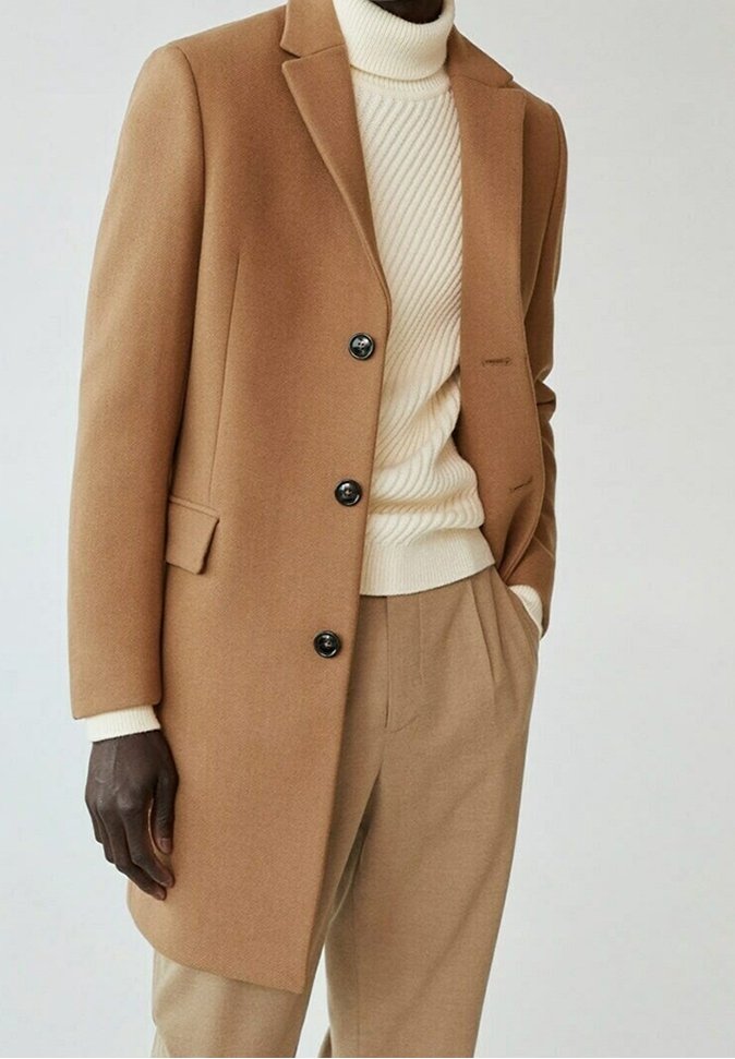 Kamelfarvet uldfrakke med klippet krave, tre sorte knapper og sidelommer, parret med en cremefarvet ribbet turtleneck og beige bukser.