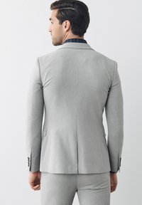 Giacca di suit grigia chiara con tessuto strutturato, con spacco singolo sul retro e tre bottoni scuri su ciascun polsino, abbinata a una camicia a scacchi sotto.