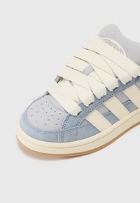 Zilā zamšādas sporta apavi ar off-white gumijas zoli, plašām strukturētām šņorēm, perforētu purngali un trim kontrastējošām sānu svītrām.