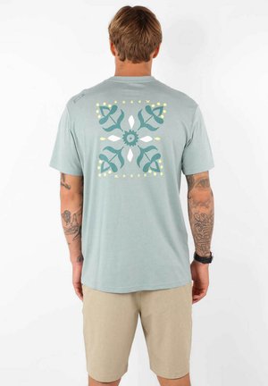 T-shirt bleu clair avec un motif graphique floral en vert, blanc et jaune au dos, assorti à un short beige. Tissu doux, manches courtes.