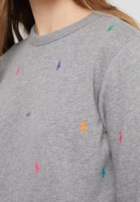 Primer plano de un suéter gris de cuello redondo con logotipos de jugador de polo bordados en varios colores esparcidos por la tela.