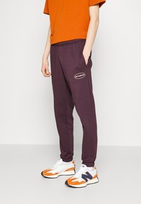 Fialové tepláky s elastickým pasem a bočními kapsami, s bílou nášivkou loga "new balance" na levé noze.