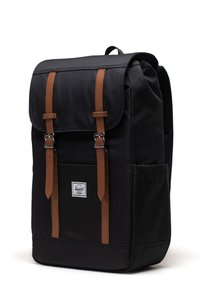 Sac à dos en tissu noir avec des sangles en cuir brun et des éléments en métal argenté, comportant une poche avant et un patch logo en bas à l'avant.