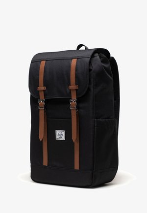 Svart ryggsäck med två bruna läderremmar med spännen och ett litet vitt märkeslappar på framfickan, enkel vit bakgrund.
