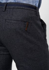 Tmavé kalhoty s vzorem a texturovanou tkaninou, s páskem v pase s modrými pruhy a malým hnědým detailem kapsy na zadní straně.