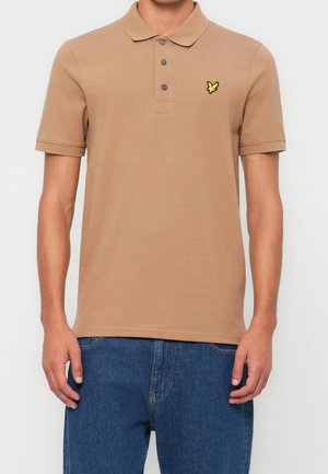 Person iført en beige kortærmet polo med tre knapper og en lille gul fuglelogo på venstre bryst, kombineret med blå jeans.