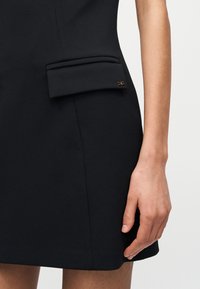 Χέρι γυναίκας ακουμπισμένο δίπλα σε μαύρο φόρεμα με πλαϊνή τσέπη με καπάκι και μικρό λογότυπο με λεπτομέρειες σε χρυσό χρώμα.