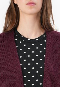 Burgundský pletený cardigan přes černou košili s bílými puntíky, který se vyznačuje texturovanou tkaninou a kulatým výstřihem.
